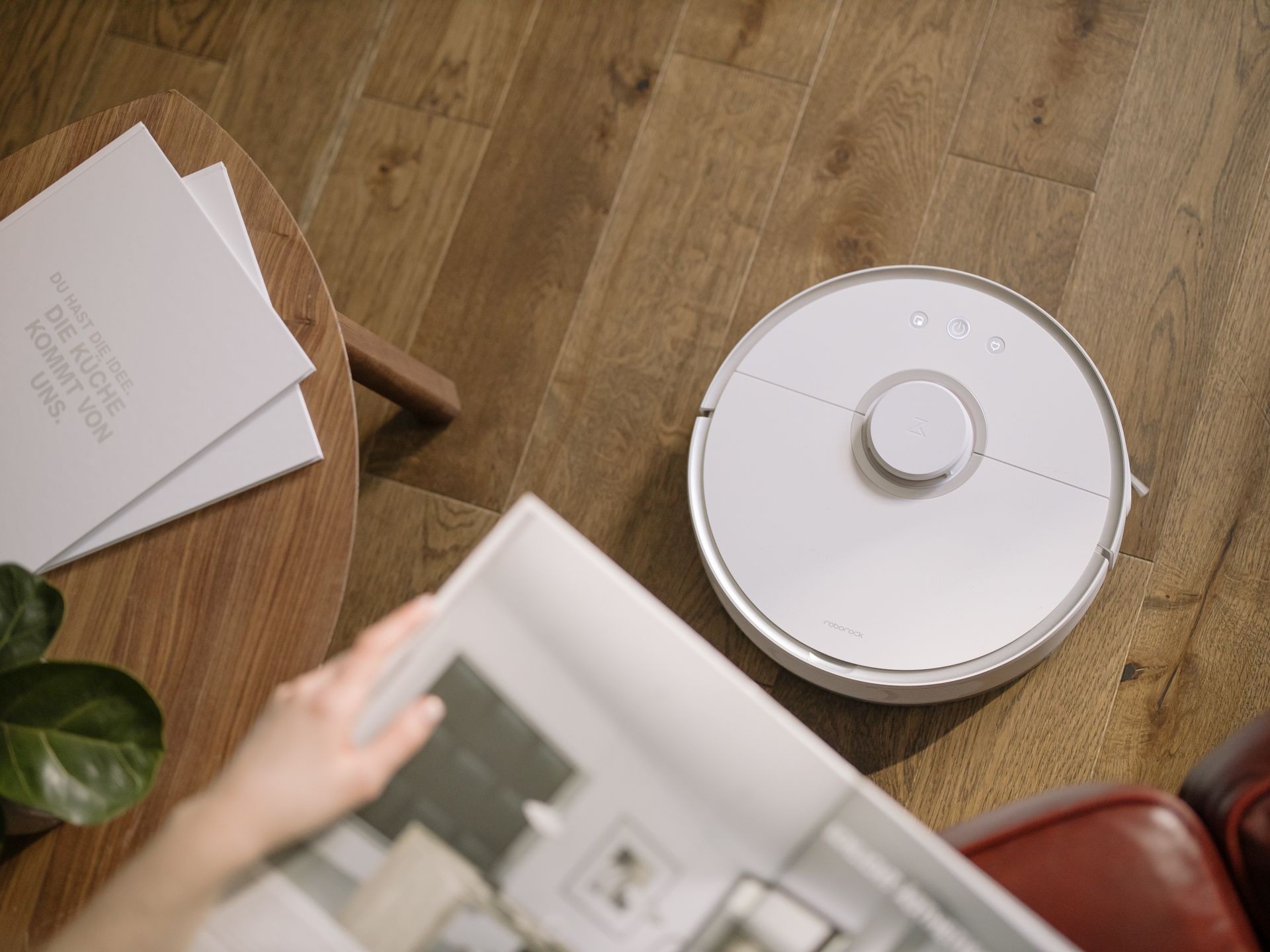 Robot vacuum cleaner on a wooden floor next to a table with open booklet and plant.